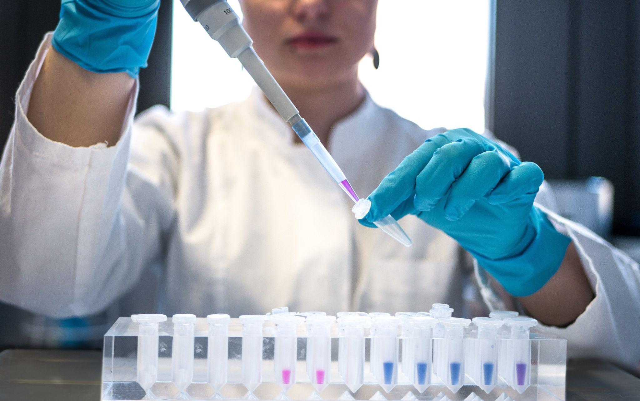 Front view of lab technician in a white coat adding some reagent to a small tube. 