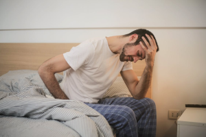 A man sits on the edge of a bed holding his head and lower back, appearing to be in pain or discomfort.