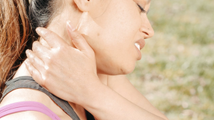 A woman gently rubs her neck, appearing to soothe muscle tension outdoors.