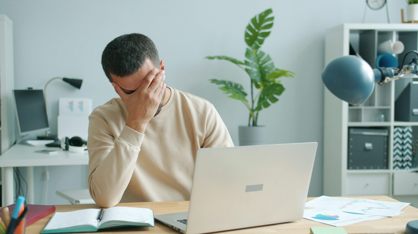 a person holding his forehead while sitting at a desk with a laptop, notebook and documents